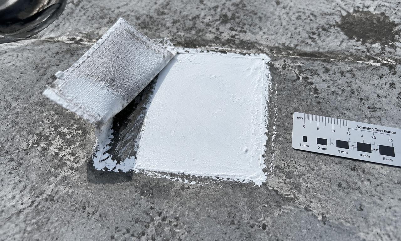 A small square test patch of white coating on a weathered gray roof, with a fabric strip partly pulled up beside a test gauge